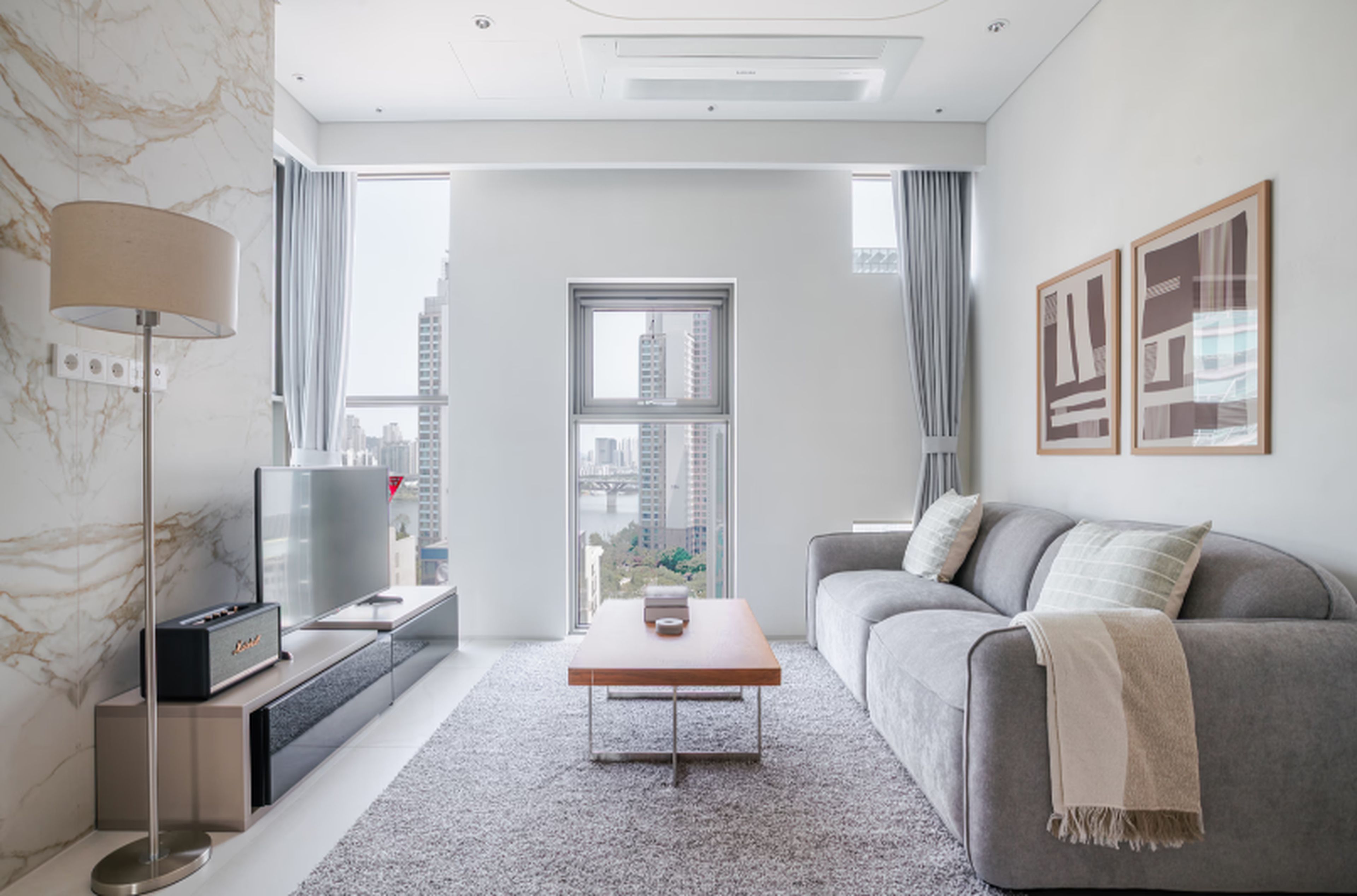 Una sala de estar moderna con un sofá gris, una mesa de centro, un televisor y vistas a la ciudad a través de amplios ventanales.