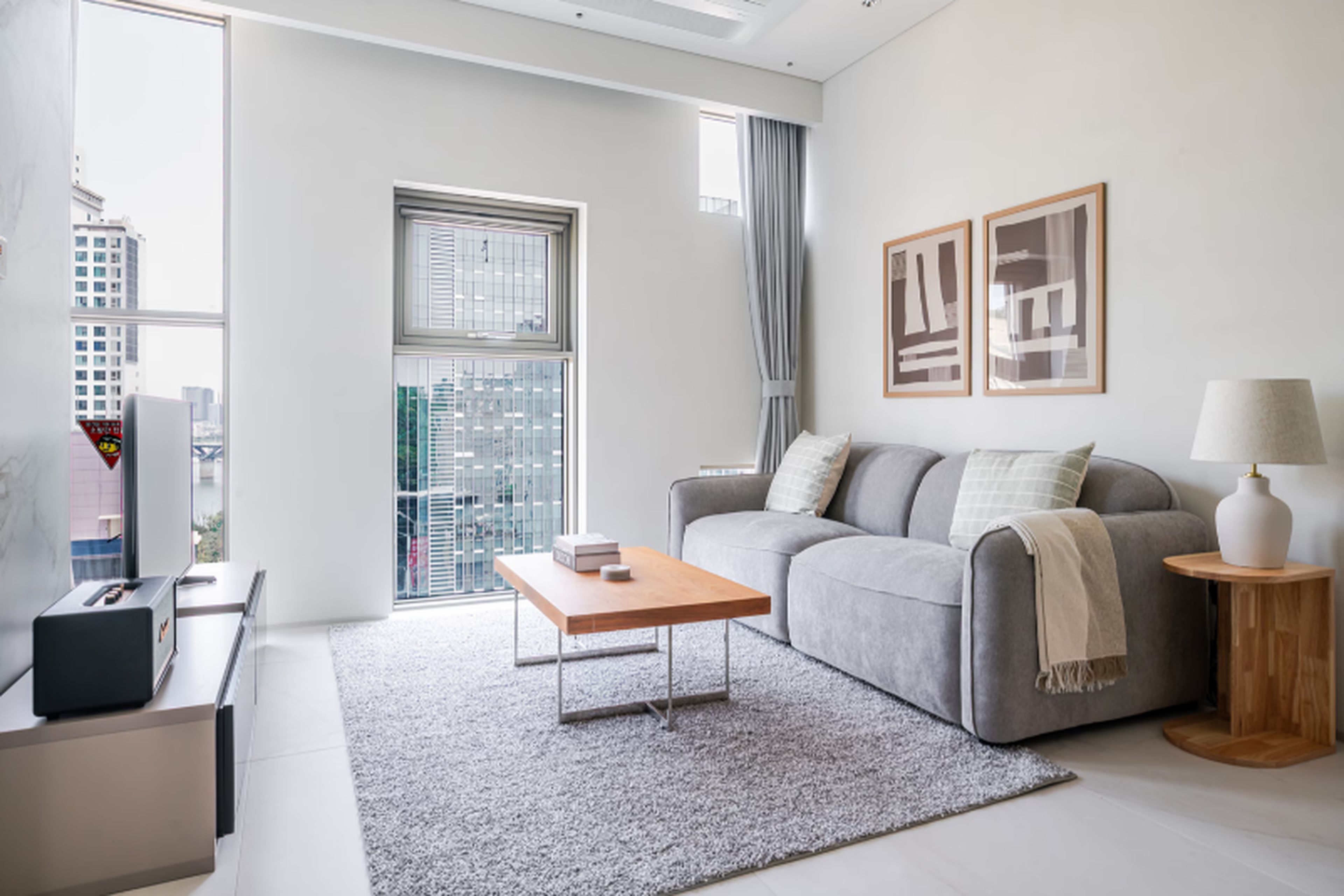 A modern living room with a grey sofa, a coffee table, and a city view through large windows.