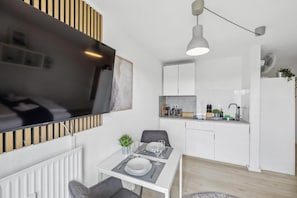 A modern kitchen with white cabinets, a dining area with a table set for two, and a TV mounted on the wall. - Go2Cozy - Study & Work Apartments (Kaiserslautern)