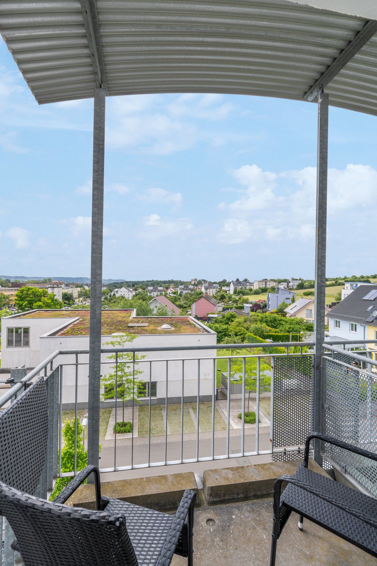Un balcón con vista a una zona residencial, con barandilla metálica y dos sillas de mimbre.