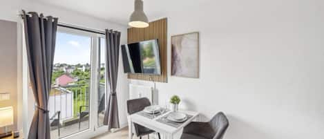 A modern dining area with a table set for two, a television mounted on the wall, and a view of the outdoors through large windows.