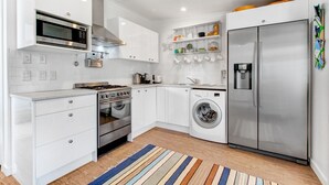 Cocina compacta con armarios blancos, refrigerador de acero inoxidable, horno empotrado, microondas, lavadora y cafetera.