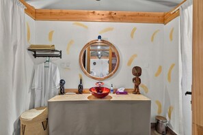 Casa de banho com teto de madeira, espelho redondo, lavatório vermelho, porta-toalhas e uma pequena figura de madeira.