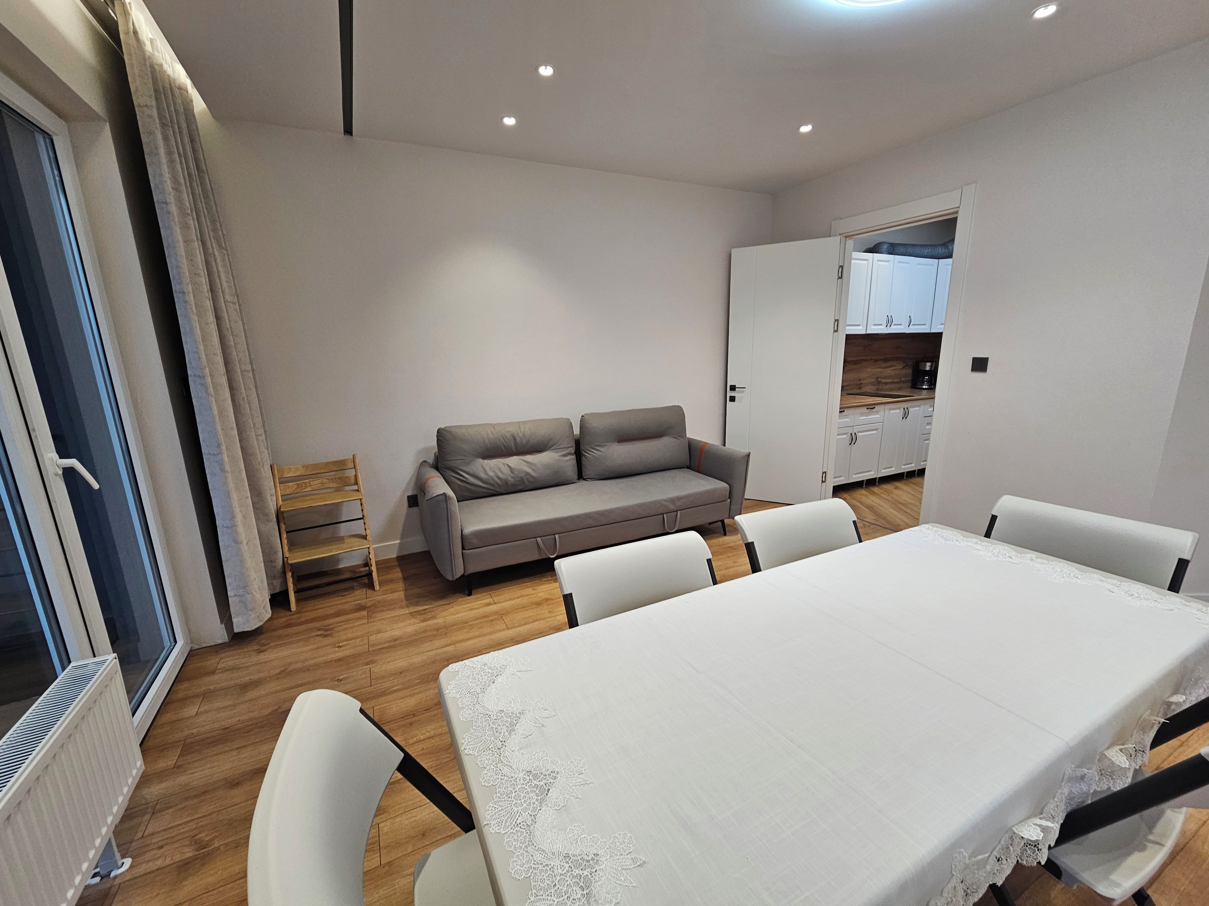 A modern living room with a sofa, dining table, and chairs. There is a kitchen area visible through an open door.