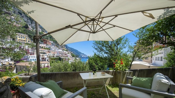 An outdoor seating area with a table and chairs, a large umbrella, and a scenic view of a hillside town. - Villa Deli (Positano)