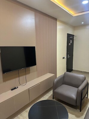 A modern living room with a flat-screen TV mounted on the wall, a grey sofa, a small round table, and a door with a handle.