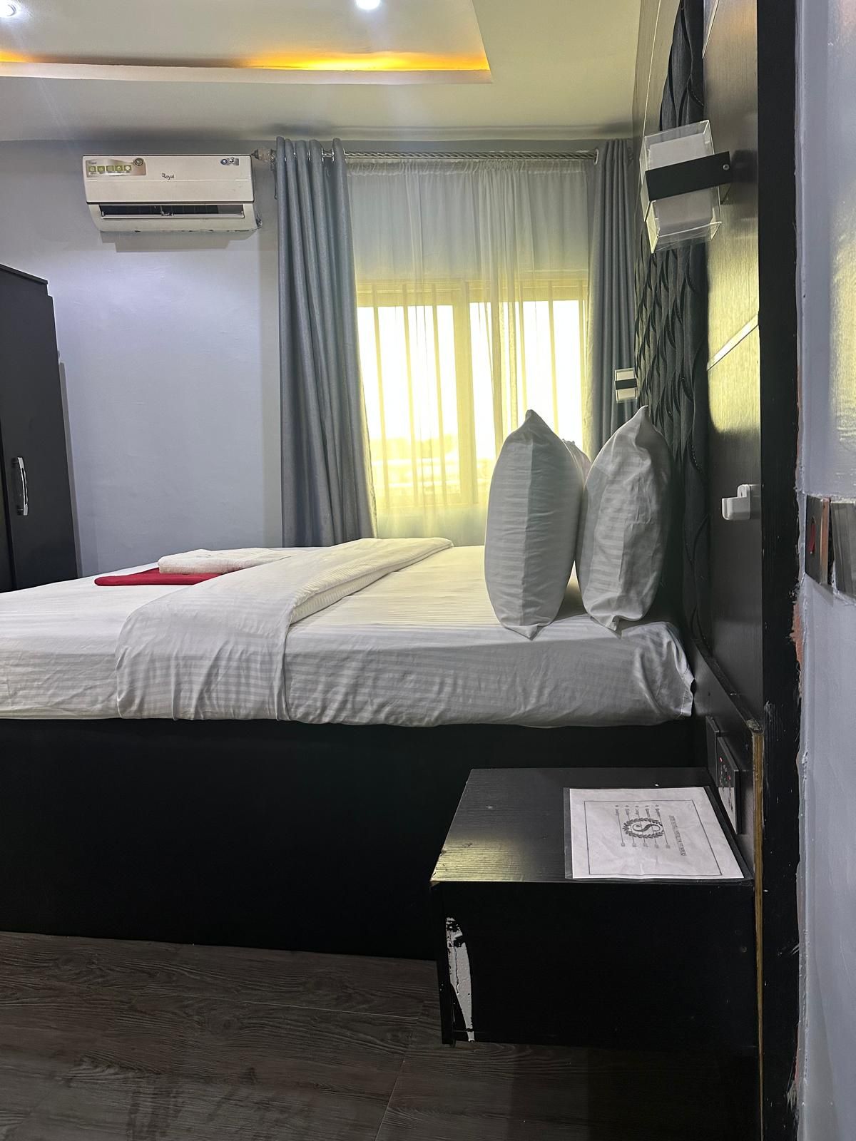 Habitación de hotel con cama, mesita de noche, unidad de aire acondicionado y ventana con cortinas.