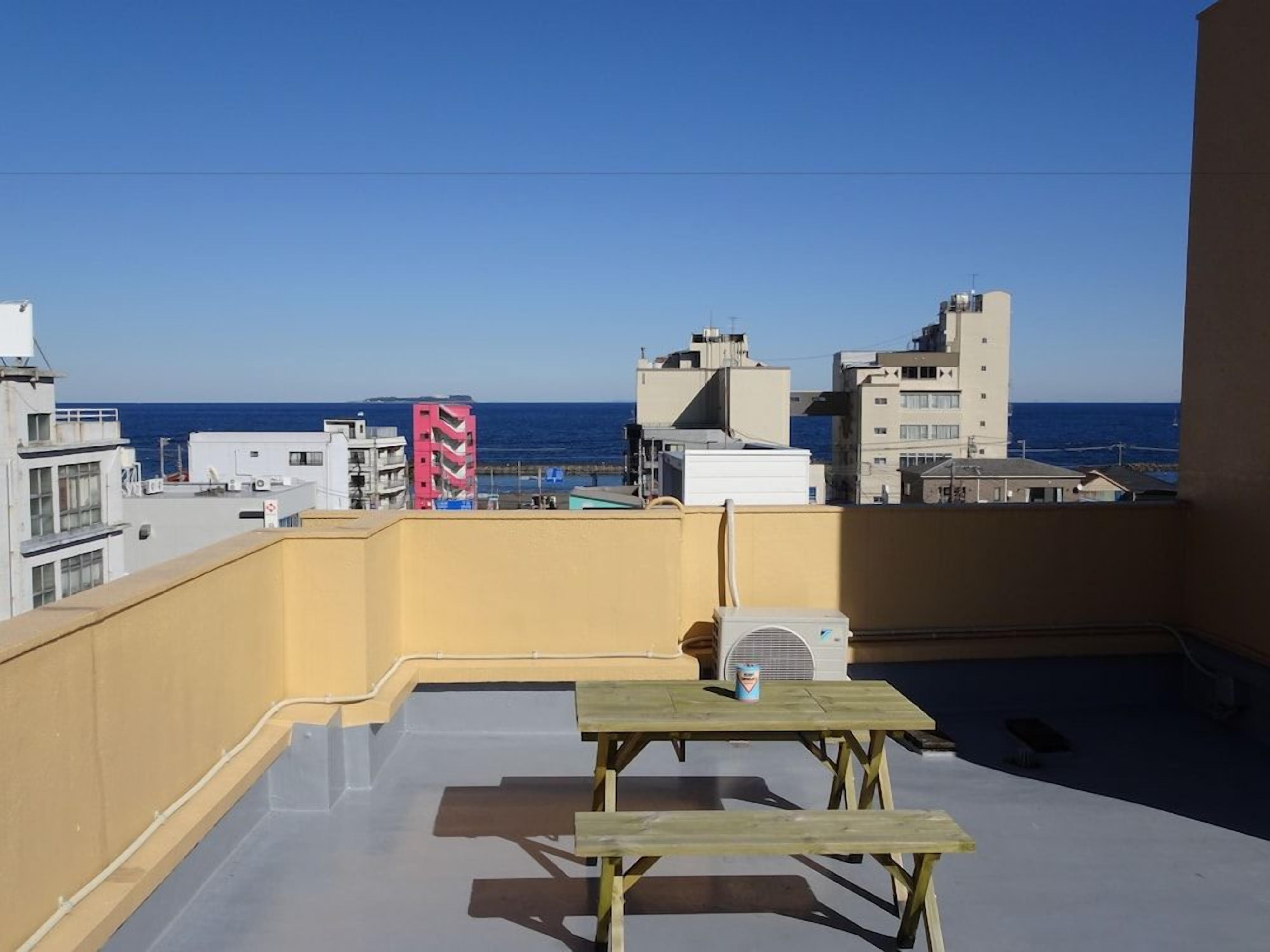 A rooftop terrace with a view of the sea, featuring a table and chairs, and a building with a yellow facade in the background.