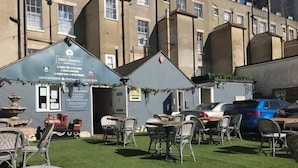 Un área de estar al aire libre con mesas y sillas, un pequeño edificio con la inscripción 'THREE CHAMNEYS' y un coche estacionado.