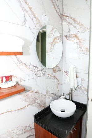 Un bany amb una paret de marbre, un mirall rodó, un lavabo blanc i una prestatgeria de fusta.