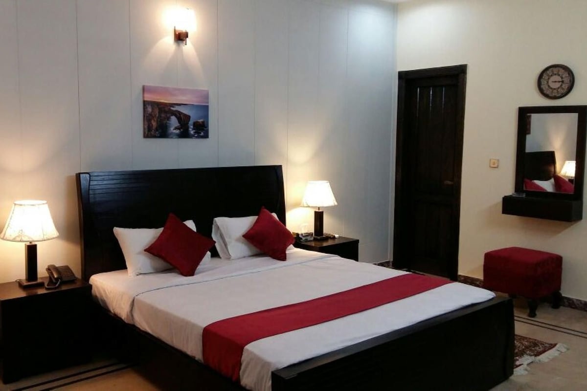 A hotel room with a bed, two bedside lamps, a nightstand, a red chair, a red ottoman, a wall clock, and a framed picture.