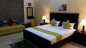 A hotel room with a bed, a sofa, a nightstand, a lamp, and a wall clock. - Serene Residence (Islamabad)