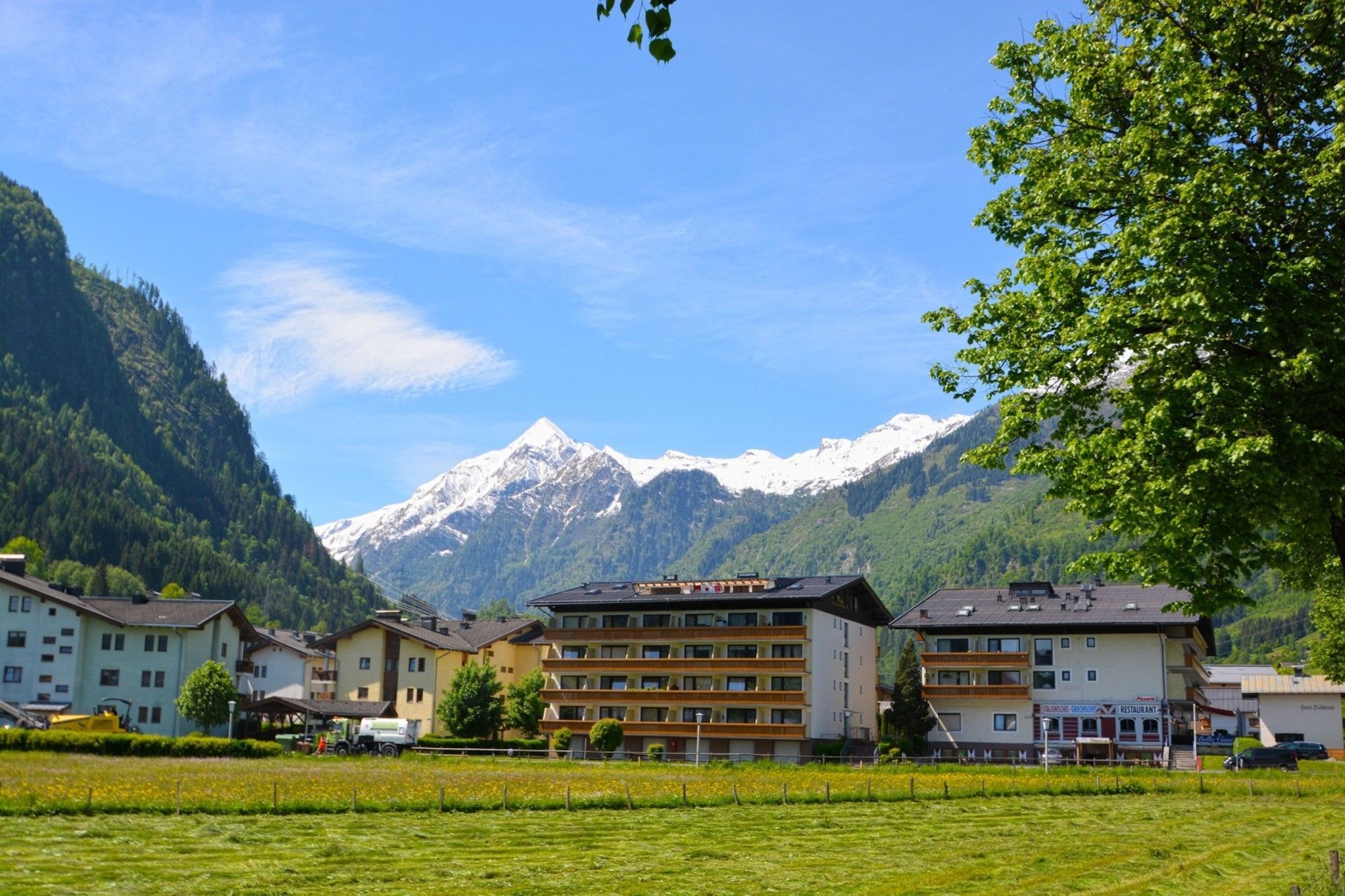 A hotel with multiple floors and balconies, set against a backdrop of snow-capped mountains and lush greenery.