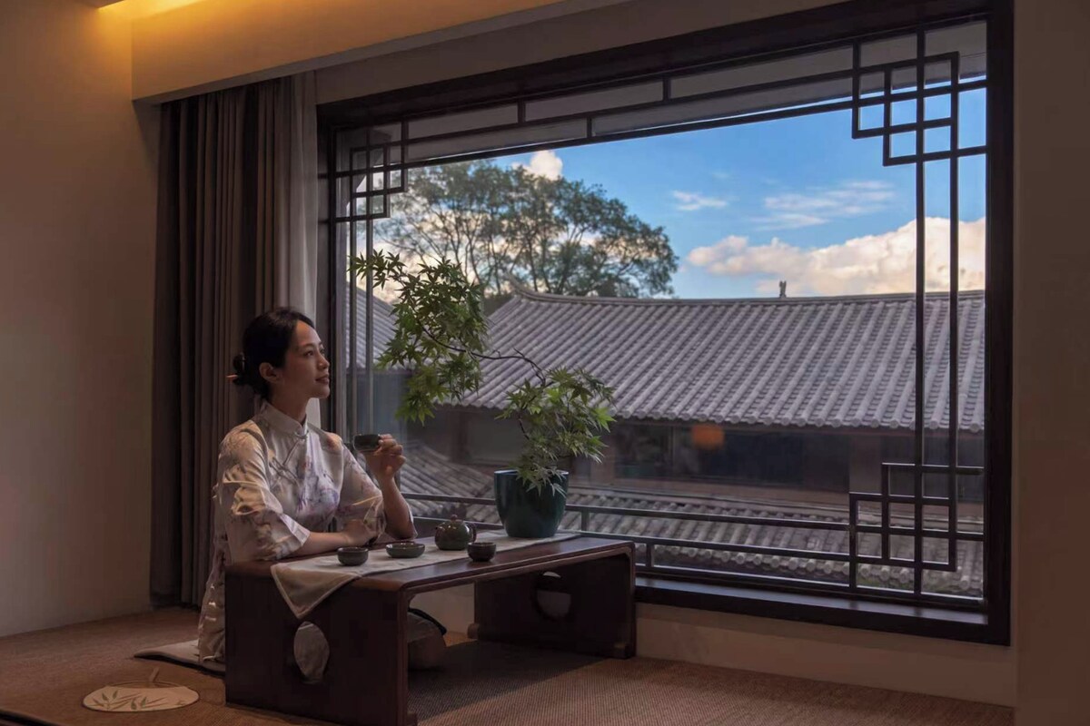 Una habitación con un área de estar tradicional japonesa con esteras de tatami, un juego de té y vistas a un edificio tradicional a través de una ventana con celosías.