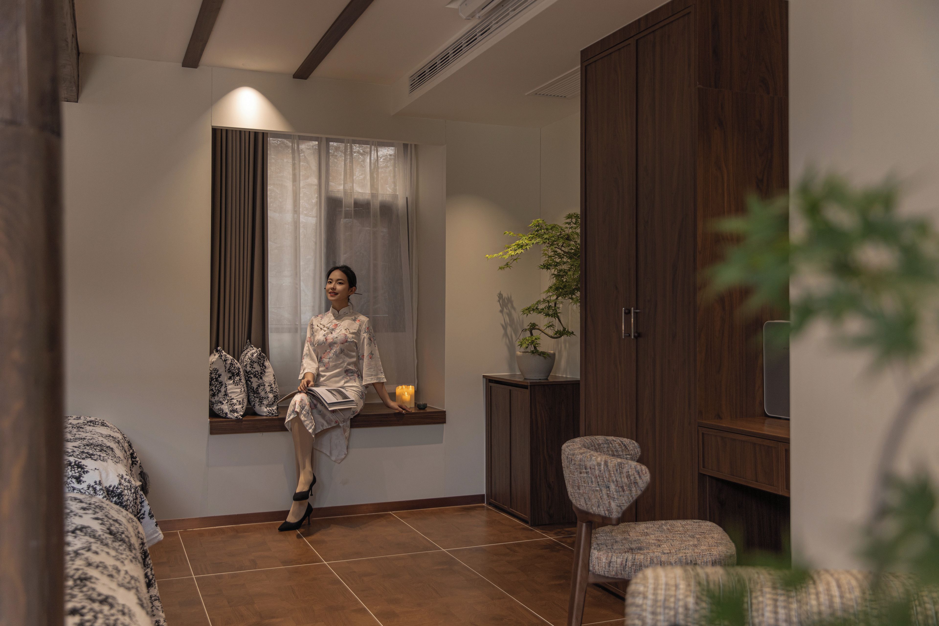 A person seated by a window with curtains, a potted plant, and a cabinet in a room with a tiled floor and a sofa.
