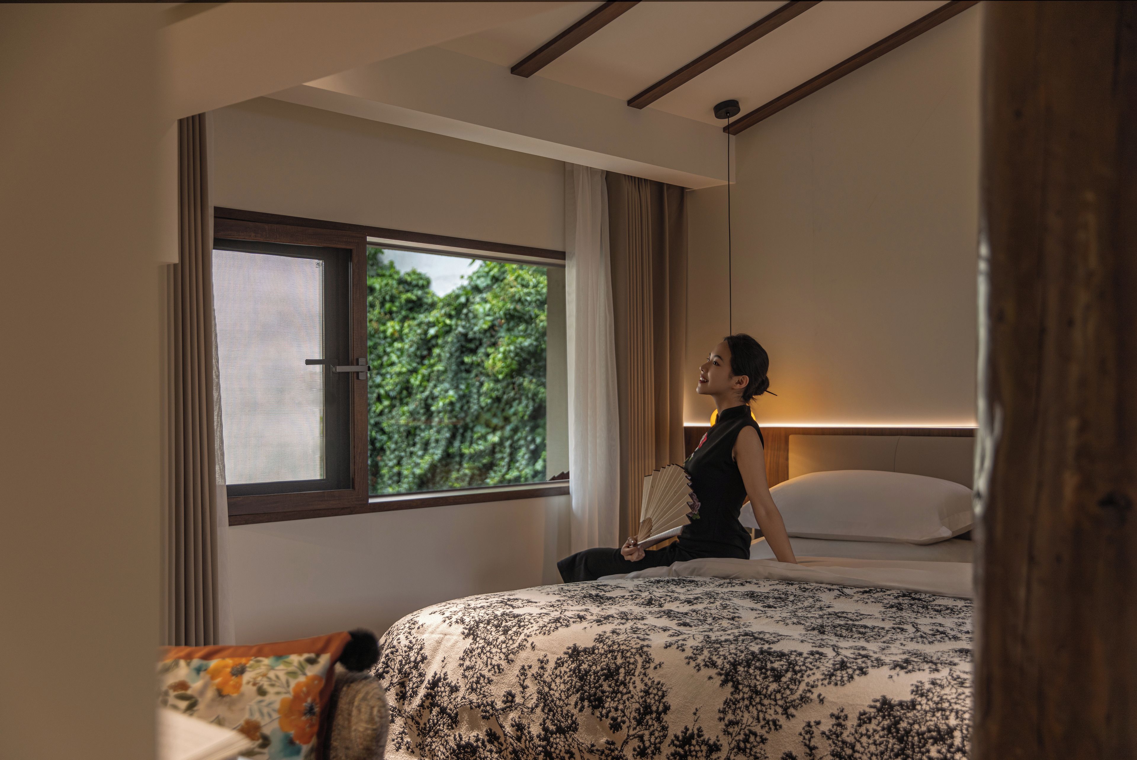 A hotel room with a large bed, a window with a view of greenery, and a bedside table with a lamp.