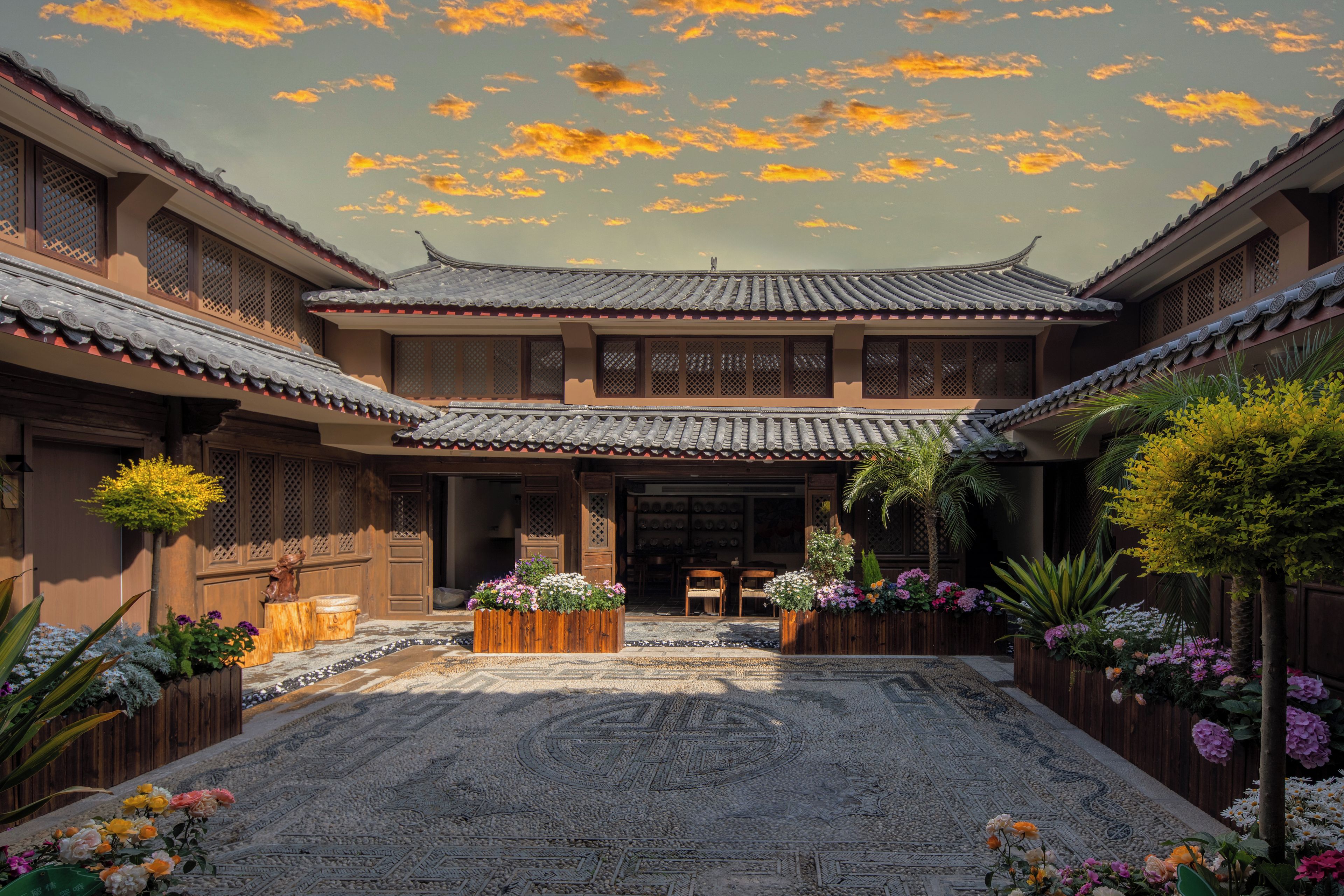 A traditional courtyard with tiled roofs, wooden structures, and a central seating area surrounded by potted plants and flowers.