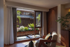 Una habitación con puerta de vidrio corrediza que da a un jardín exterior, una silla de madera con cojín y una mesita con un libro.