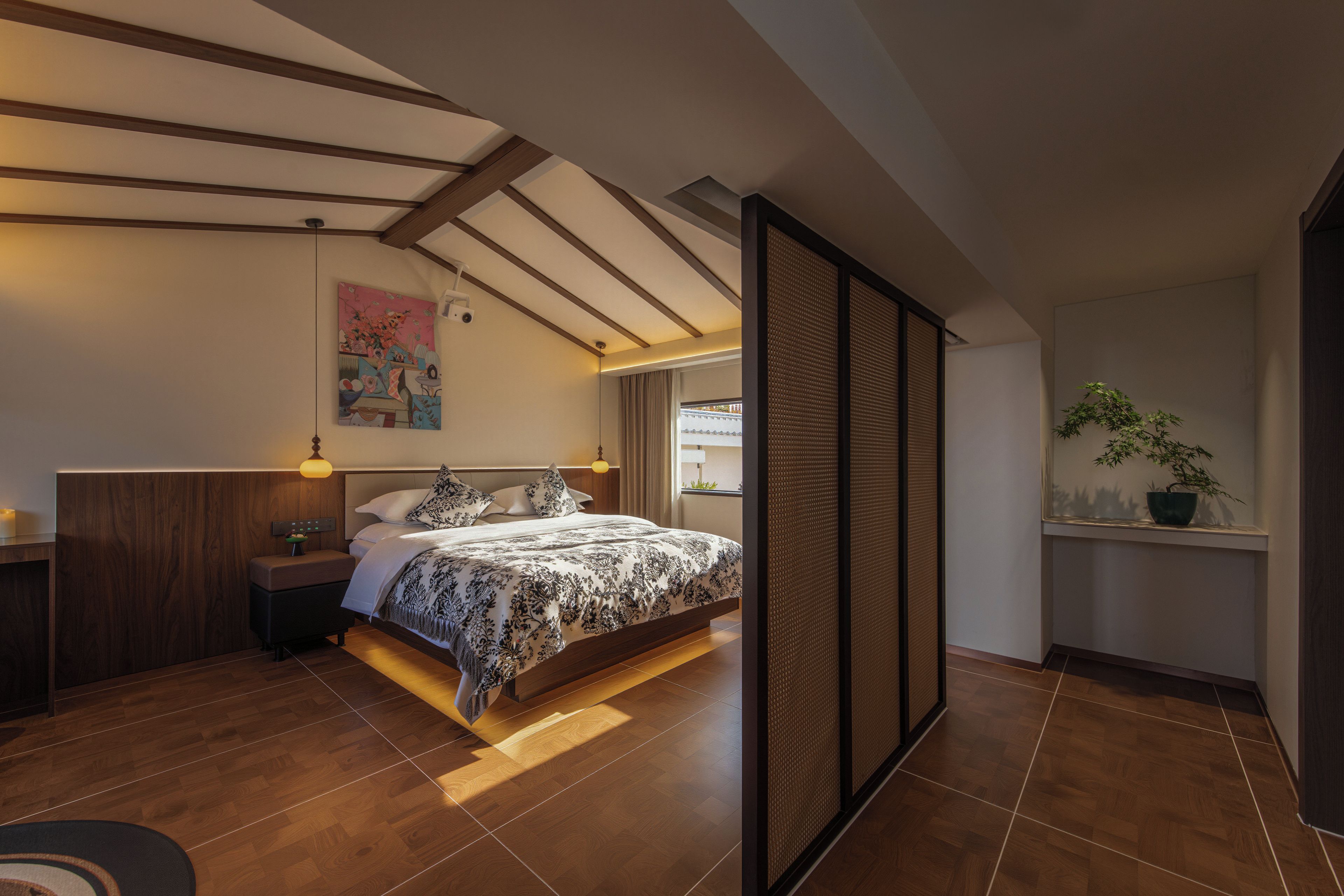 A modern bedroom with a large bed, wooden flooring, a nightstand, a bedside lamp, and a potted plant.