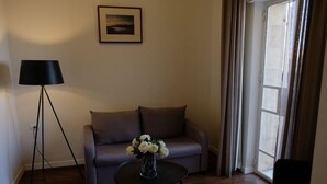 Ein Zimmer mit einer Couch, einem kleinen Tisch mit Blumenvase, einer Stehlampe und einem Fenster mit Vorhängen.