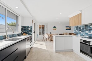 A modern kitchen with a central island, a dining area, and a view through a glass door. - "Leanda Sea Breeze"- Short walk to beaches & shops (Port Macquarie)