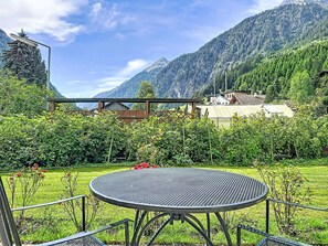 Ein runder Metalltisch mit Metallgestell, umgeben von einem Metallzaun, in einer grünen Umgebung mit roten Blumen und einer Bergkulisse.
