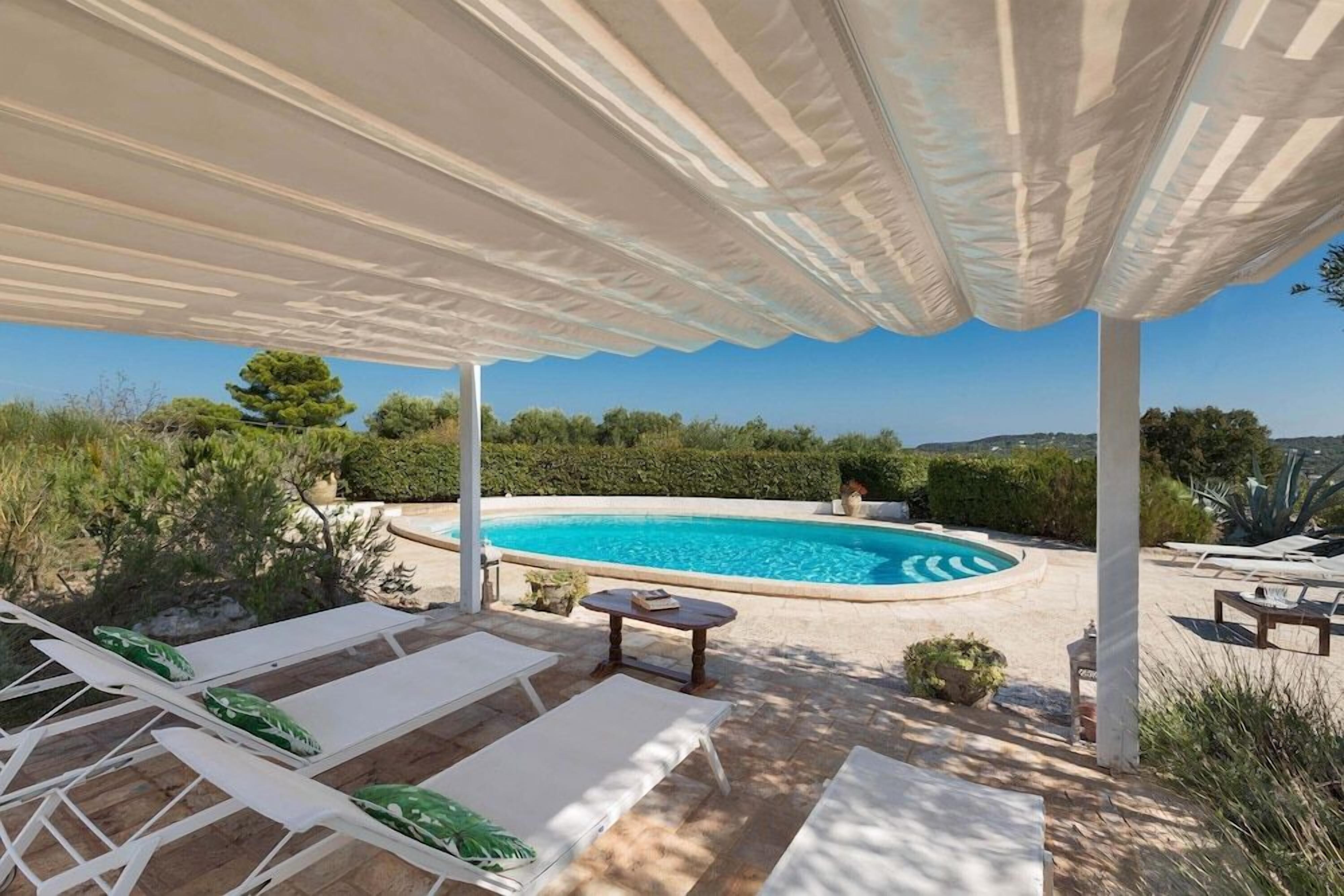 A covered outdoor area with a swimming pool, lounge chairs, and a view of the surrounding landscape.