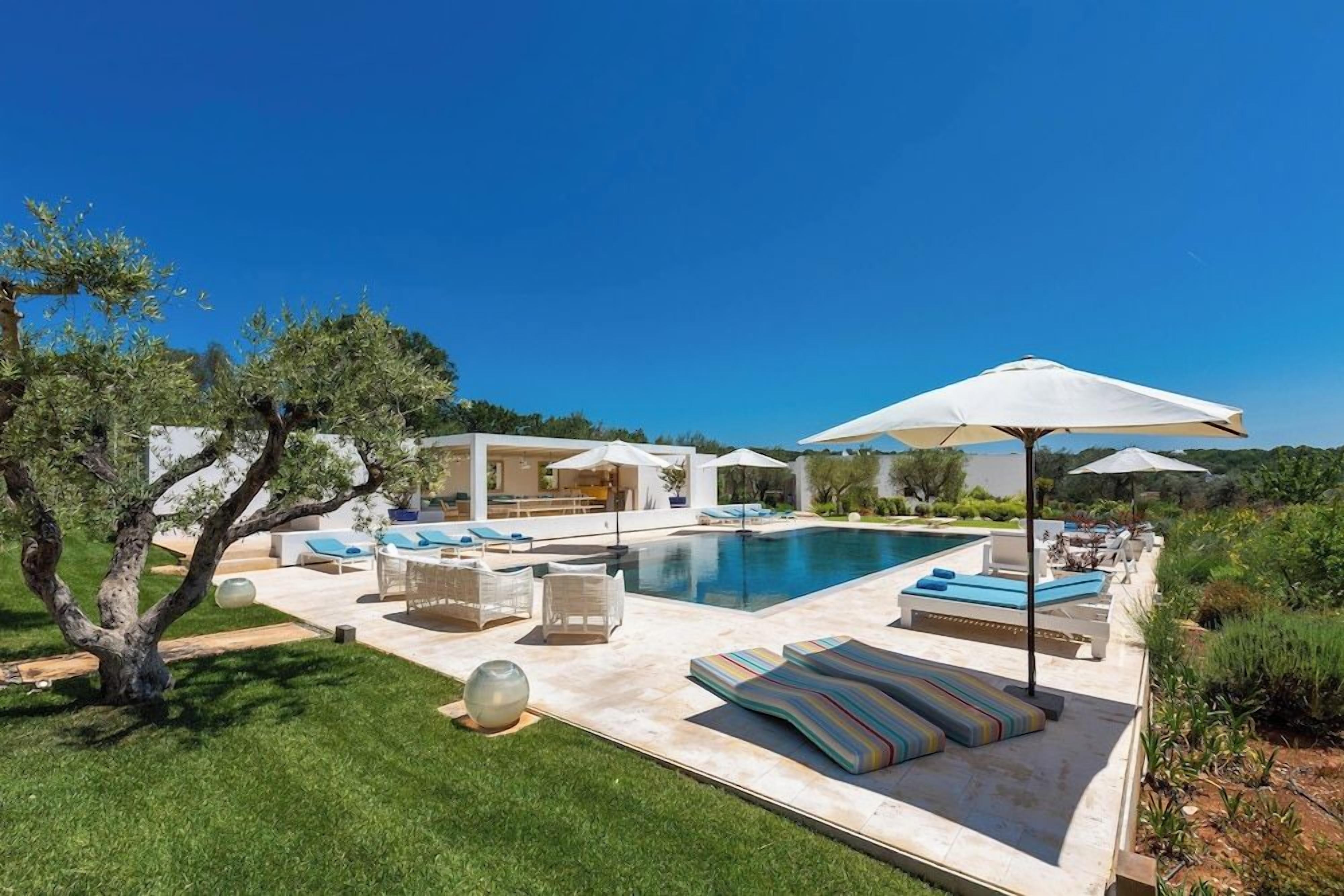 A pool area with lounge chairs and umbrellas, surrounded by greenery and a clear sky.
