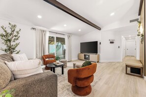 Ein modernes Wohnzimmer mit Sofa, Sessel und Hocker, einem Flachbildfernseher und einer Glastür, die zu einem Außenbereich führt.