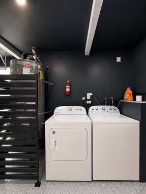 A laundry room with two white Whirlpool washers, a fire extinguisher, a metal rack with papers, and a bottle of detergent. - Modern & Spacious Anaheim Home 7 Min to Disneyland Game Room Theater Sleeps 14 (Anaheim)