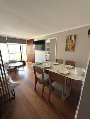 Un comedor con una mesa dispuesta para cuatro, sillas de madera y un televisor montado en la pared.