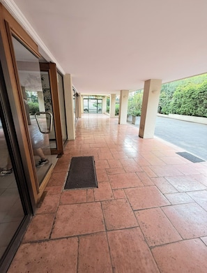 Un pasillo interior con suelo de baldosas, una entrada con puerta de cristal y vistas a una zona exterior verde.