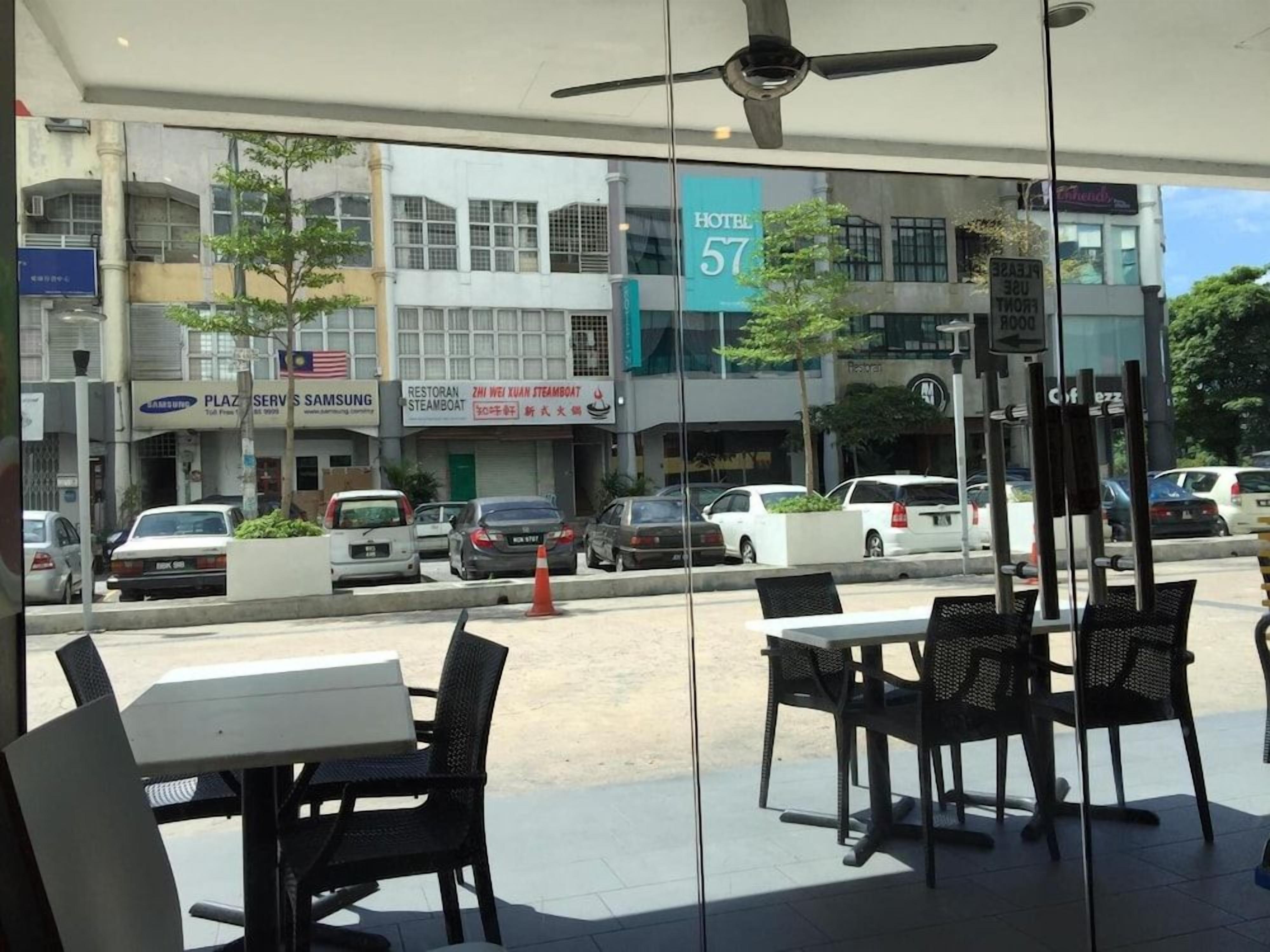 Un espace extérieur aménagé avec des tables et des chaises, une vue sur la rue où sont garées des voitures, et une enseigne d’hôtel en arrière-plan.