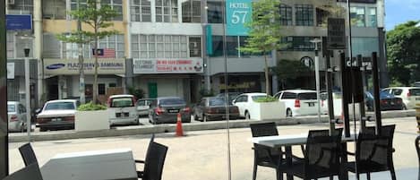 An outdoor seating area with tables and chairs, a view of the street with parked cars, and a hotel sign in the background.