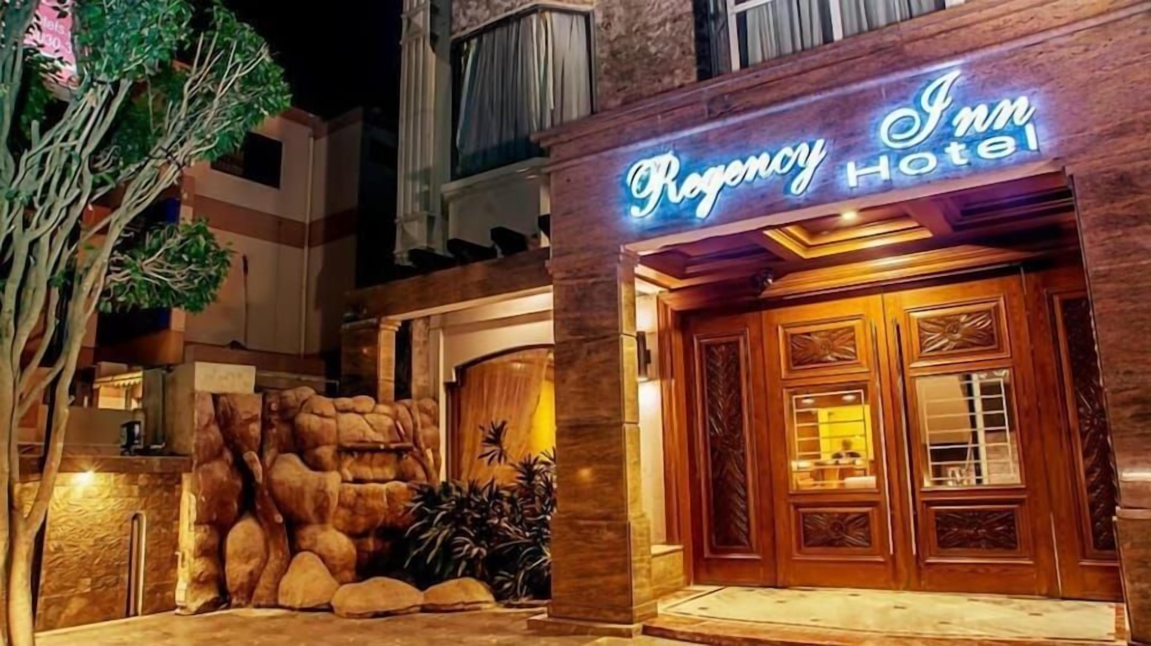 The exterior of Regency Hotel with a prominent neon sign, a stone facade, and a large wooden entrance door.