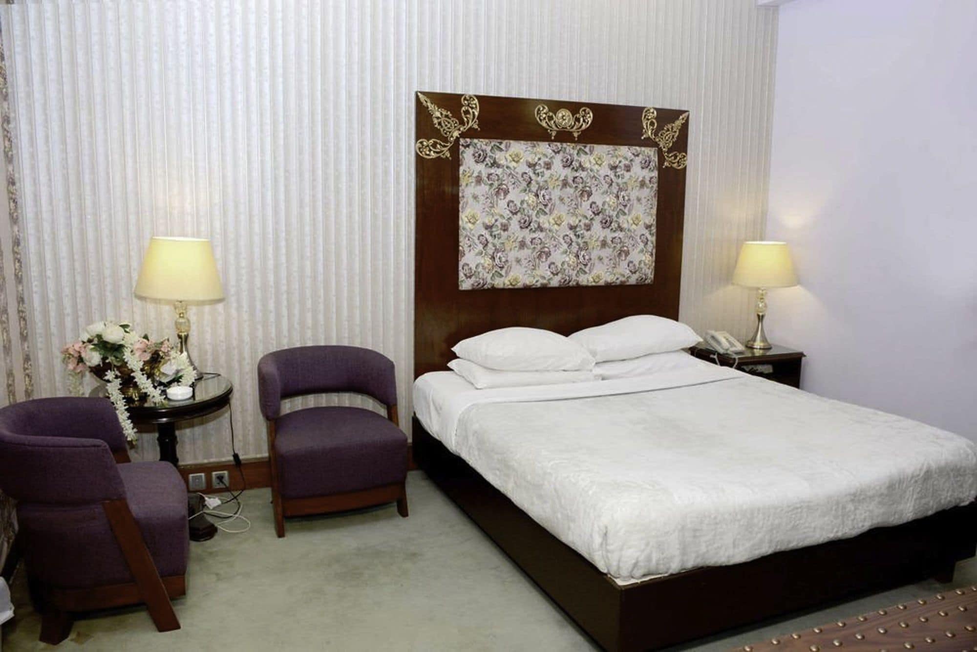 Una habitación de hotel con una cama, dos sillas, una mesita de noche con una lámpara y un cabecero estampado con motivos florales.