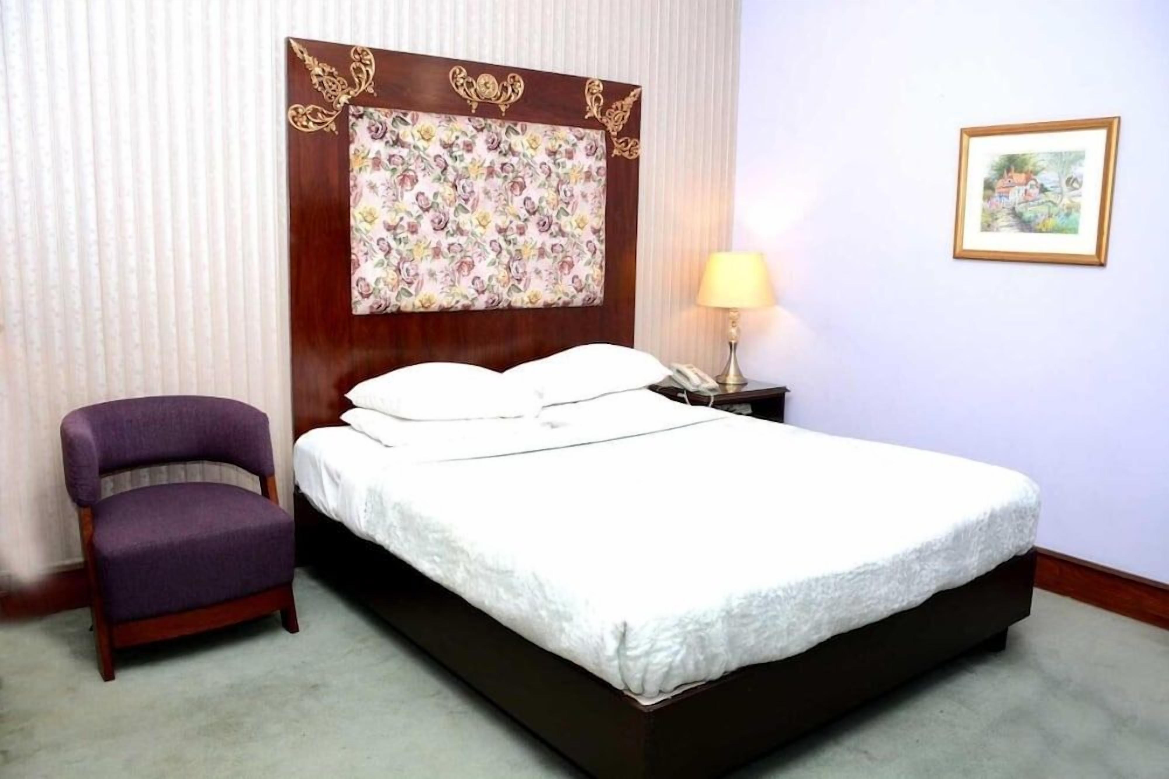 A hotel room with a bed, a chair, a nightstand, a lamp, and a framed picture on the wall.