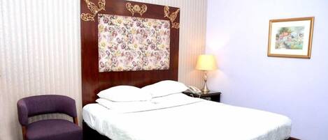 A hotel room with a bed, a chair, a nightstand, a lamp, and a framed picture on the wall.
