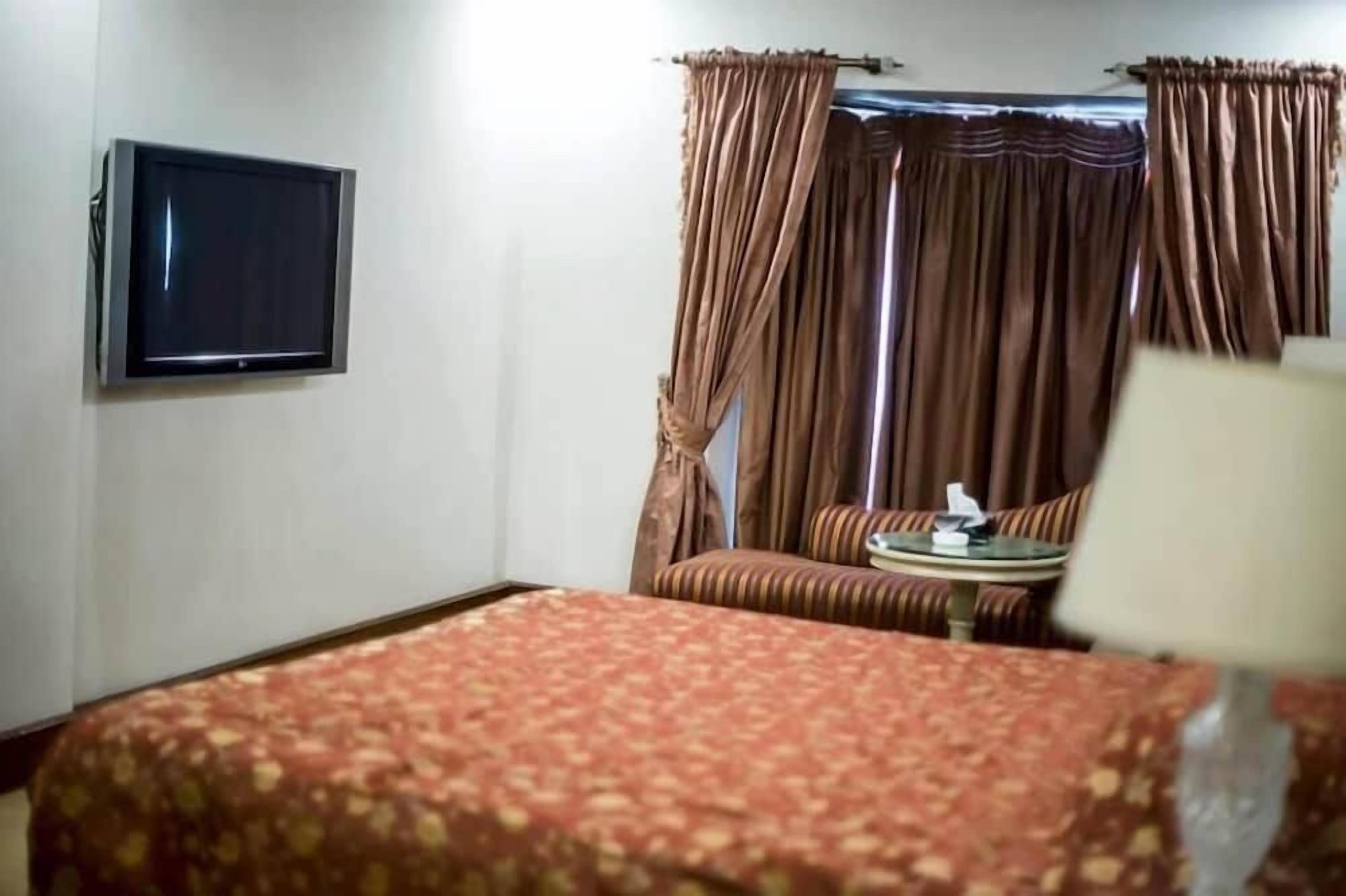 A hotel room with a bed, a flat-screen TV mounted on the wall, a small round table with a lamp, and a window with curtains.