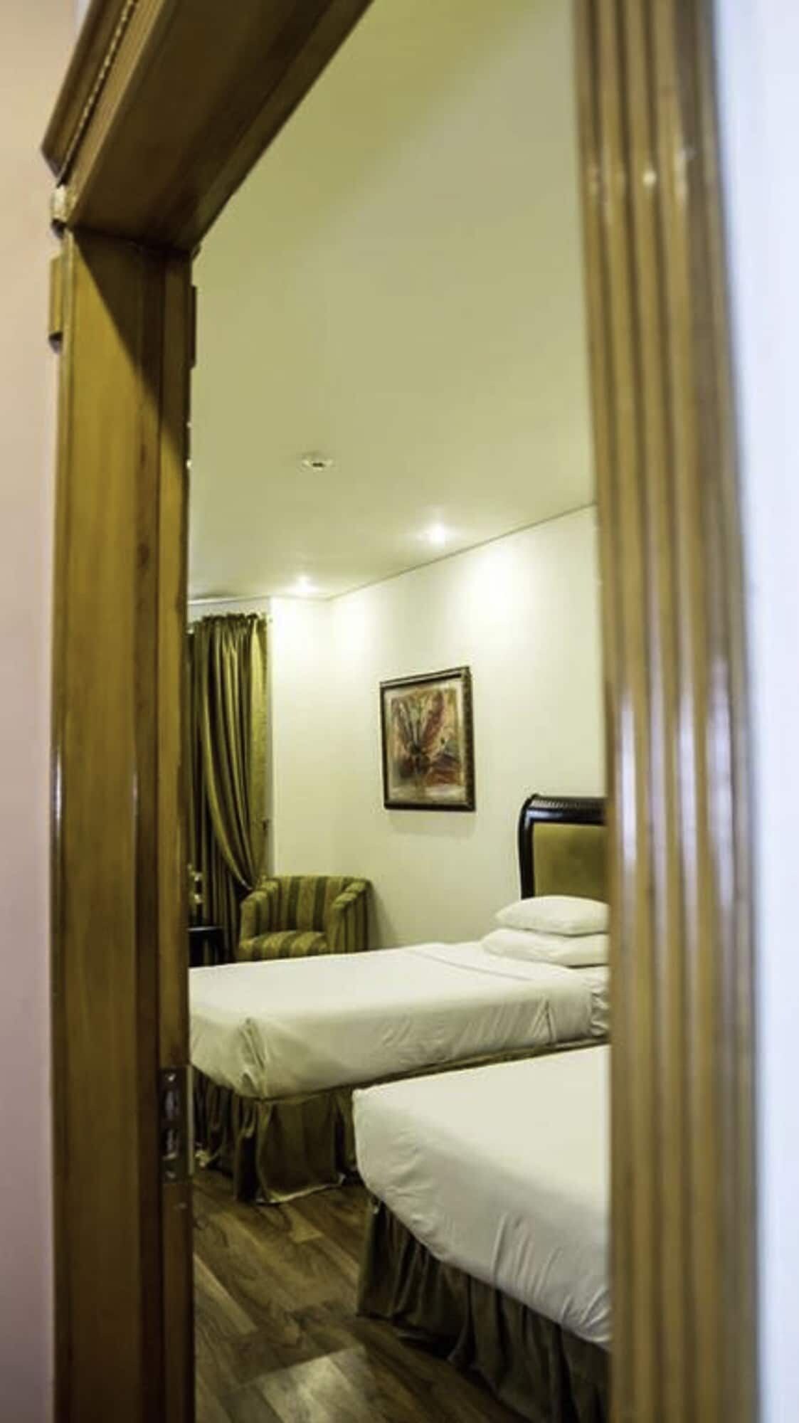 Een hotelkamer met twee bedden, een stoel, een schilderij aan de muur en een houten deuropening.