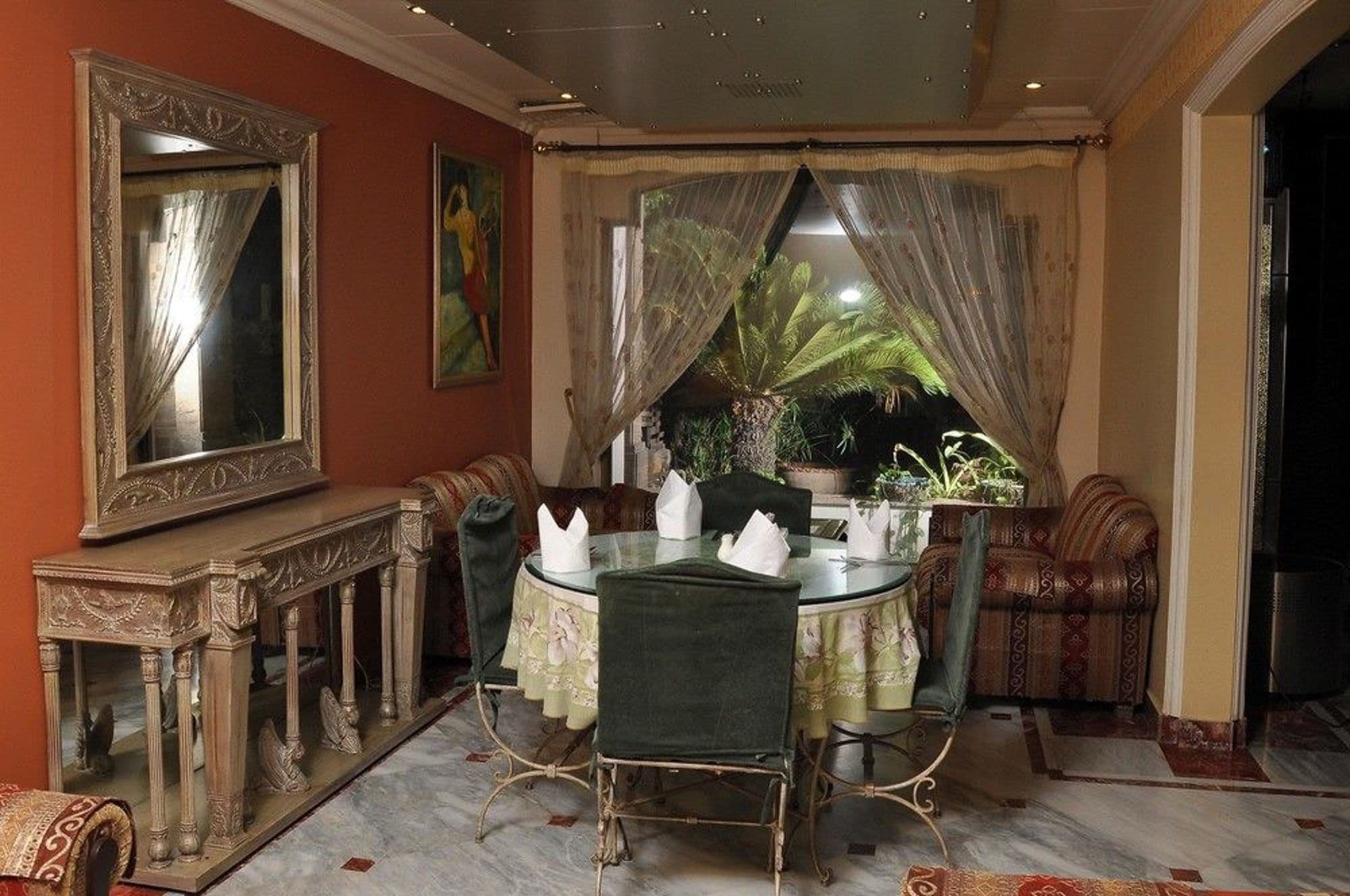 Un comedor con una mesa redonda, sillas, un espejo y vistas a un jardín a través de cortinas traslúcidas.