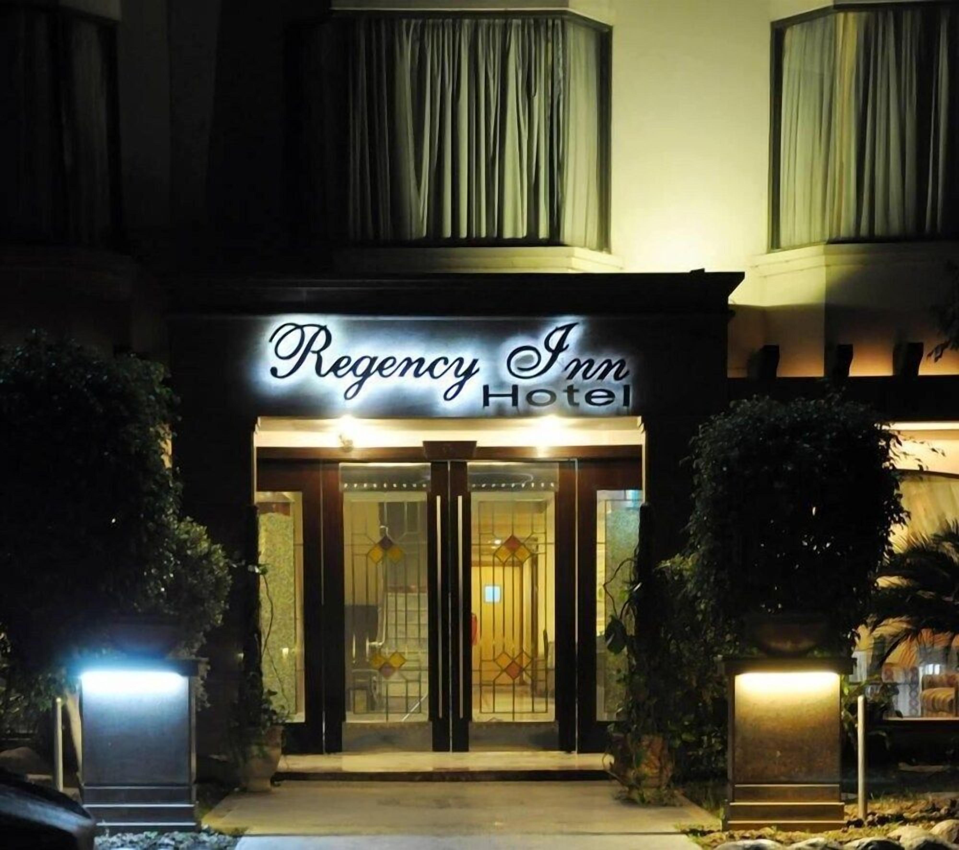 Exterior del Hotel Regency Inn con un llamativo letrero iluminado, entrada de vidrio y zona de acceso bien iluminada.