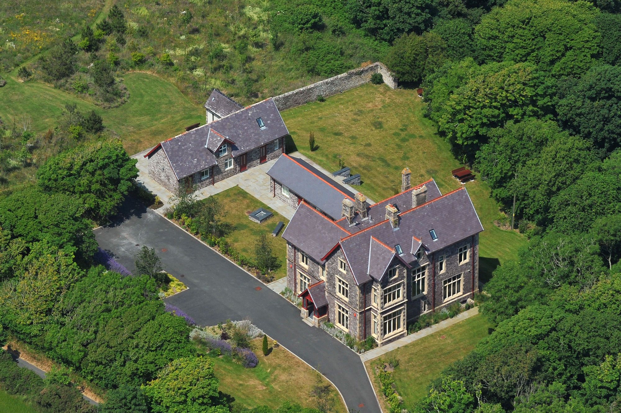 An aerial view of a large, multi-story stone building with a prominent entrance, surrounded by lush greenery and a winding road.