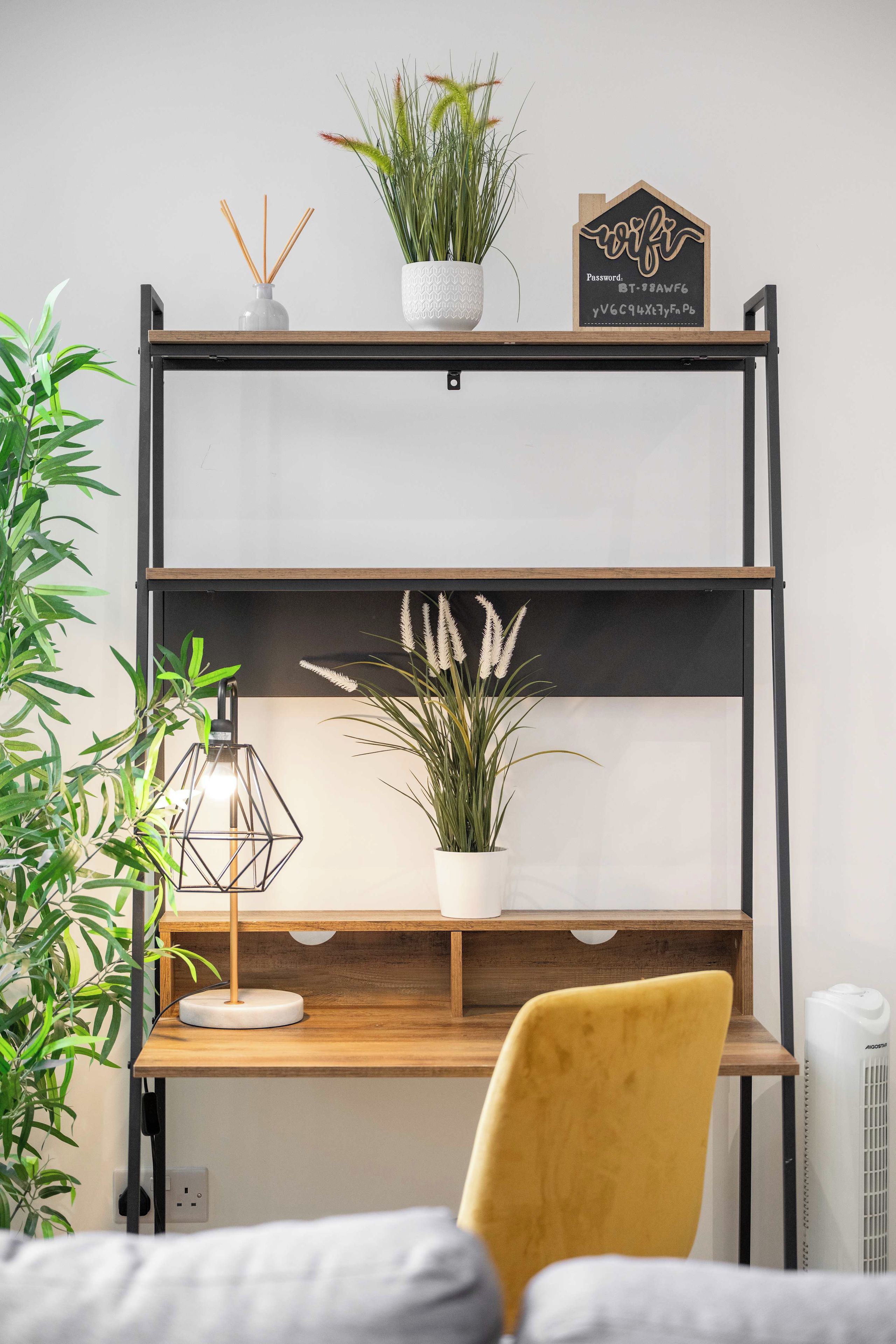 Un escritorio de madera con una silla amarilla, una planta en maceta, un cartel decorativo y una lámpara geométrica.