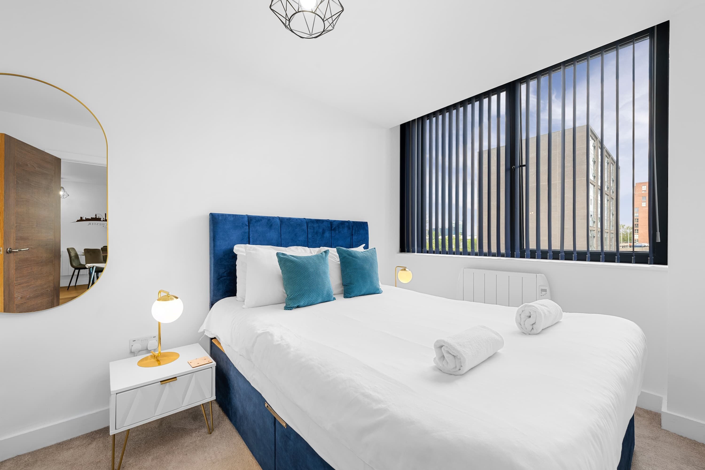 Une chambre à coucher moderne avec un grand lit, une table de chevet avec une lampe, un miroir et une fenêtre avec des stores.
