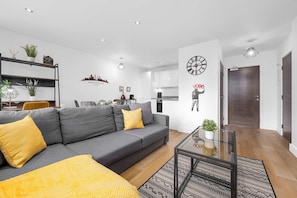 Een moderne woonkamer met een grijze bank, gele kussens, een glazen salontafel en een stadslandschap op de muur.