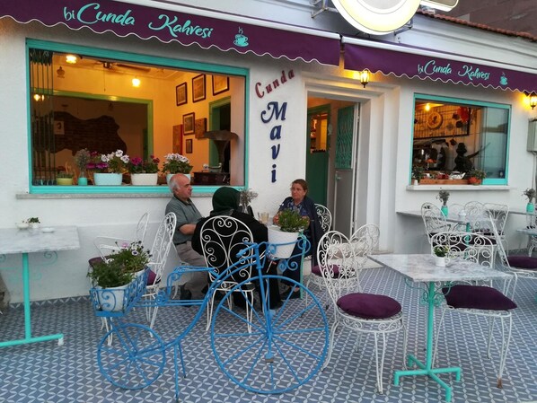 Un café al aire libre con sillas y mesas de metal, un letrero que dice 'Cunda Mavi' y plantas en macetas en el alféizar.