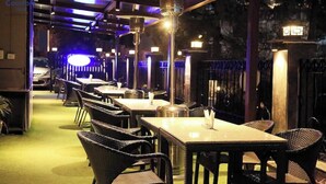 A restaurant with multiple tables and chairs, a visible trash bin, and a fence in the background. - Silver Arch Hotel (Dehradun)
