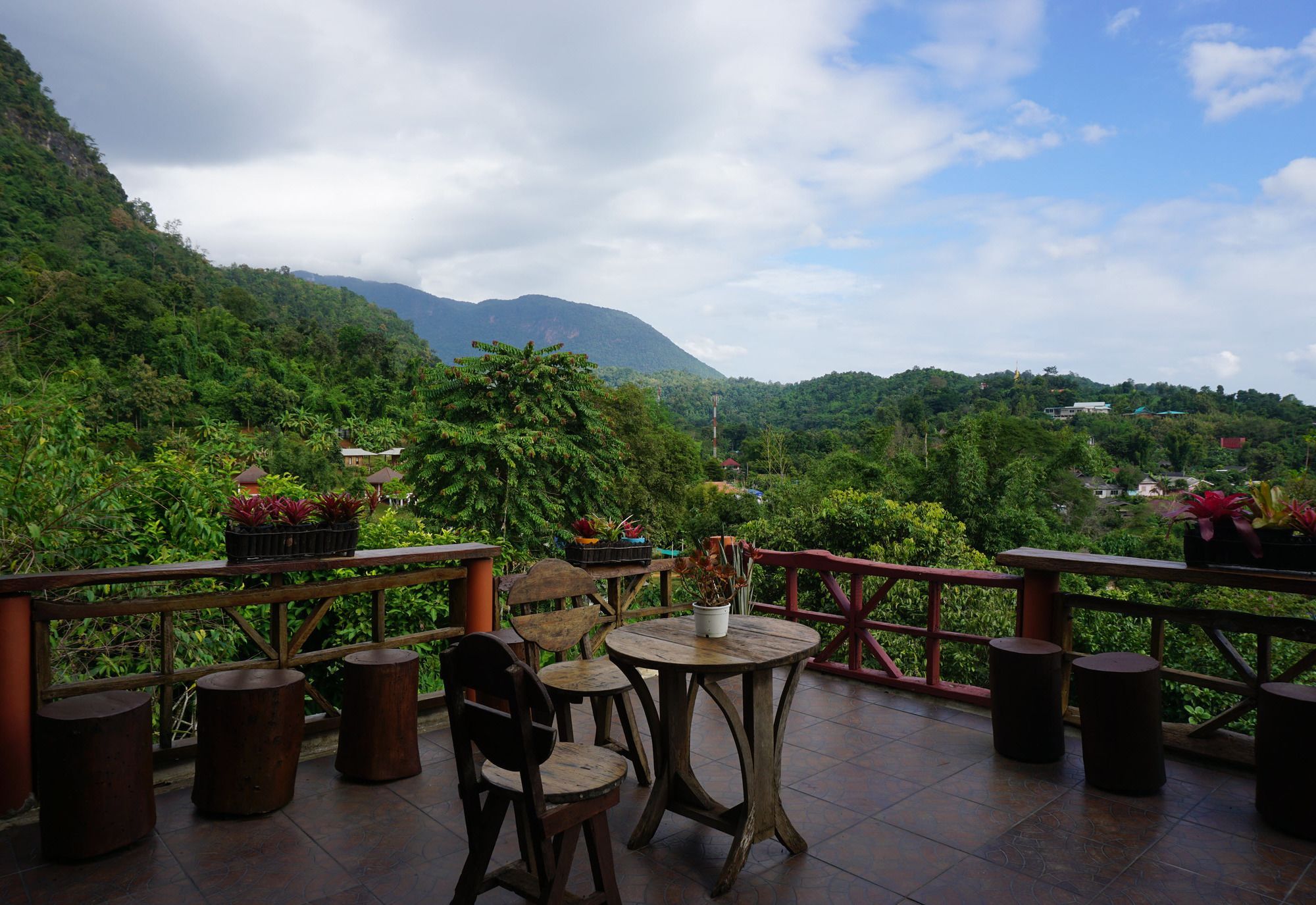 A terrace with wooden furniture and potted plants, overlooking a lush green landscape with mountains in the distance.