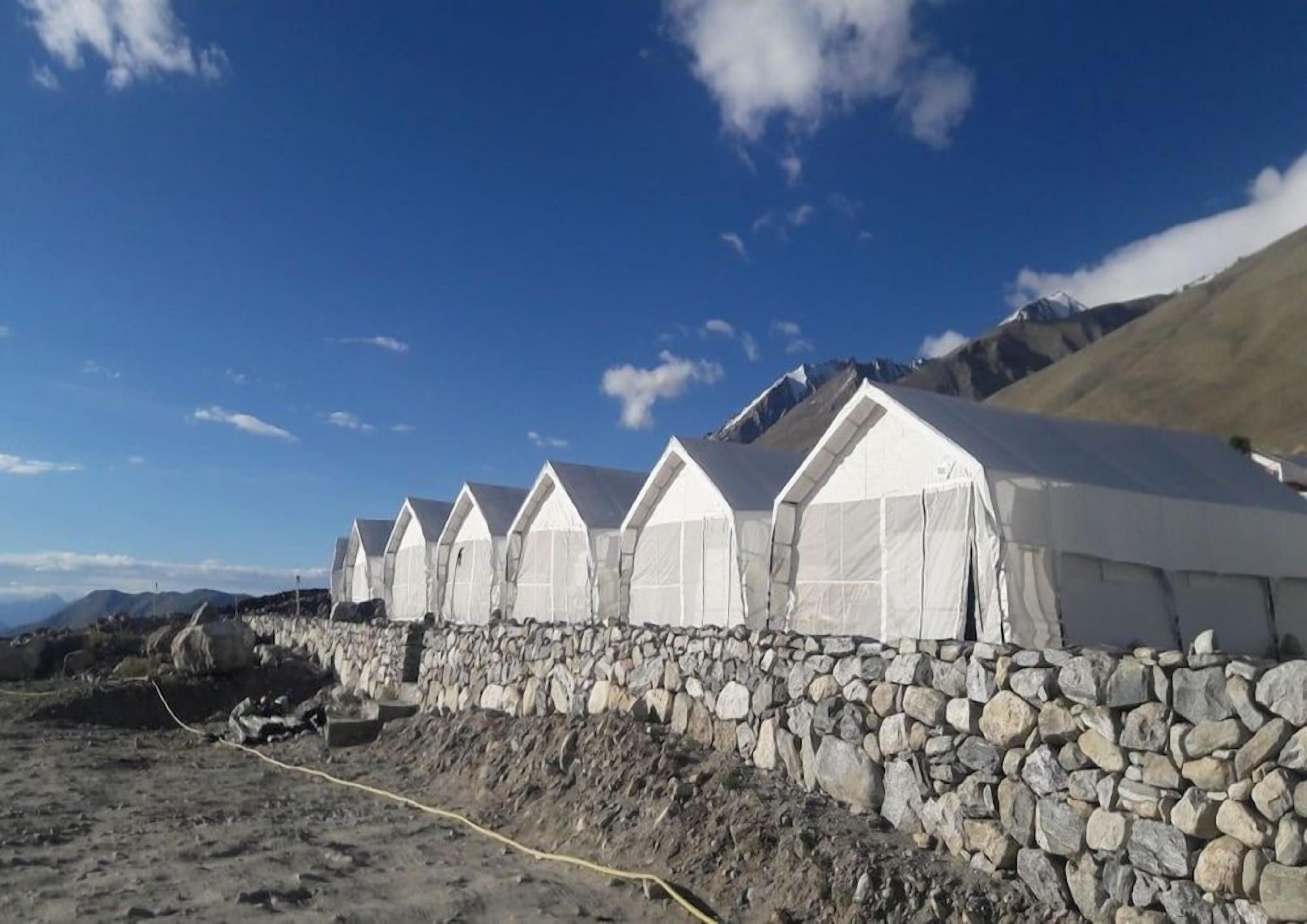 A row of white tents with pitched roofs, arranged along a stone wall, set against a mountainous backdrop.